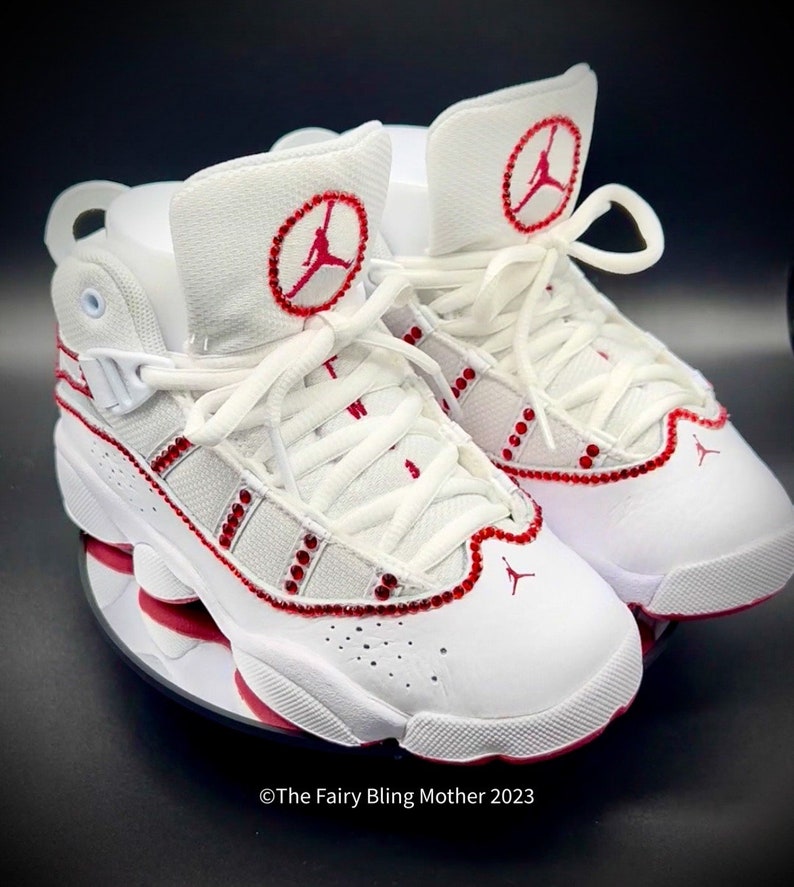 May include: White leather trainers with red accents and rhinestones. The trainers have a red Jumpman logo on the side and a red circle with a Jumpman logo on the tongue.