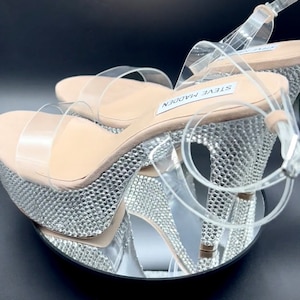 May include: Clear platform sandals with a rhinestone-covered heel and platform. The sandals have a single strap across the foot and an ankle strap. The brand name "STEVE MADDEN" is visible on the heel.