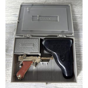 Op de afbeelding: Een vintage speelgoedpistoolset in een grijze plastic koffer. De set bevat een goudkleurig en bruin speelgoedpistool, een zwarte holster en een kleine grijze doos met het opschrift "9mm LUGER AMMUNITION". De koffer is gemarkeerd met "MARX".