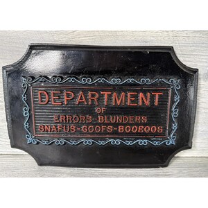 Puede incluir: Letrero rectangular negro con las palabras "DEPARTMENT OF ERRORS-BLUNDERS SNAFUS-GOOFS-BOOBOOS" en letras rojas y azules. El letrero tiene un borde decorativo y probablemente está hecho de metal.
