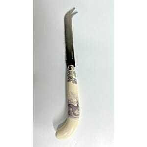 May include: A cheese knife with a stainless steel blade and a cream-colored handle. The handle features a rabbit illustration and floral accents. The knife has a serrated edge and a forked tip. The brand name "Spode" is visible on the blade.