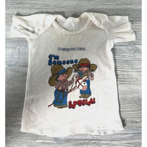 May include: Off-white baby onesie with a graphic of two cartoon cowboys and a horse. The text "Dogpatch USA" and "I'm Someone Special!" are printed on the front. The onesie has short sleeves and a round neckline.