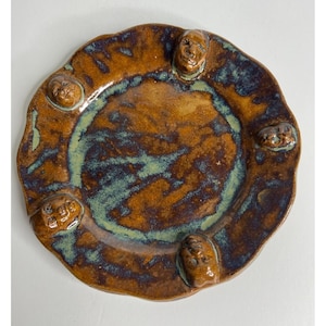 May include: A handmade ceramic plate with a unique design. The plate features a brown and blue glaze with a wavy rim. Six small, sculpted faces are evenly spaced around the edge of the plate, adding a whimsical touch to this decorative piece.