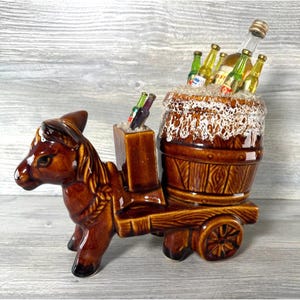 May include: A brown ceramic donkey pulling a wooden cart with a barrel. The barrel is filled with ice and miniature liquor bottles. The donkey has a detailed mane and harness, and the cart has a rustic design.