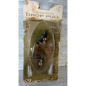 Może przedstawiać: Zabytkowy uchwyt do szafki na kartonowym podkładzie. Okucia mają ciemny brązowy kolor z ozdobnymi detalami. Na opakowaniu widnieje napis "Quality Cabinet Accessories DROP PULL" i "NATIONAL LOCK". Zawiera dwie srebrne śruby.