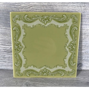 Puede incluir: Azulejo de cerámica cuadrado verde claro con un borde decorativo y ornamentado en un tono más claro de verde. El borde presenta diseños florales y de hojas en espiral, que enmarcan un cuadrado central liso.
