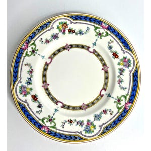Pode incluir: Um prato de porcelana branca com um design floral. O prato apresenta uma borda azul e amarela com um padrão repetitivo. O centro do prato é branco, com um design floral preto e rosa ao redor da borda interna. Diâmetro de aproximadamente 20 cm.