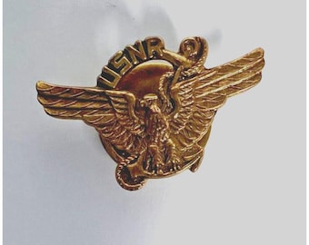 Puede incluir: Un pin USNR dorado con un águila con las alas extendidas, un emblema circular y las letras "USNR" en la parte superior. El pin tiene detalles intrincados y un acabado pulido.