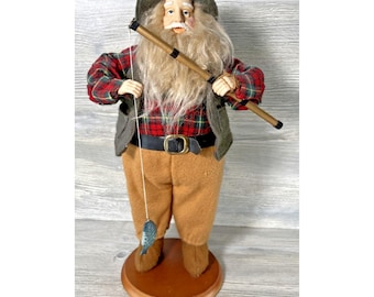 Puede incluir: Figura decorativa de un anciano vestido con una camisa a cuadros roja y negra, pantalones marrones y un chaleco verde. Sostiene una caña de pescar con un pez pequeño adjunto. La figura está sobre una base de madera.