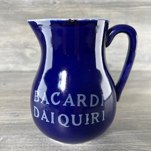 Può includere: Brocca in ceramica blu cobalto con manico curvo. Le parole "BACARDI DAIQUIRI" sono stampate in bianco sul davanti. La brocca ha una finitura lucida e un bordo leggermente scheggiato.
