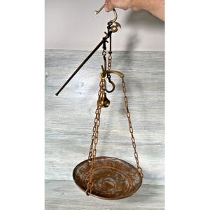 Op de afbeelding: Antieke hangende weegschaal met een koperkleurige ovale pan die aan kettingen is opgehangen. De weegschaal heeft een messing haak en een lange, zwarte arm om te meten. De weegschaal is ontworpen voor het wegen van items.