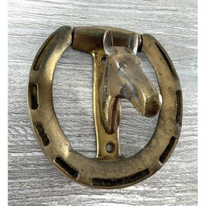 May include: A brass door knocker in the shape of a horseshoe, with a horse head design. The gold-coloured metal knocker features a detailed horse head in the centre. The horseshoe has a textured surface.
