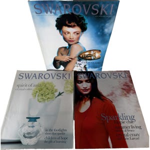 May include: Three Swarovski magazines are displayed. The top magazine cover features a woman holding a mask. The other two magazines have text that reads "spirit of asia" and "Sparkling hippie chic". The magazines are on a white background.