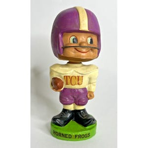 May include: A vintage TCU Horned Frogs bobblehead figurine. The figure wears a purple helmet with a yellow stripe, a cream-colored jersey with "TCU" on the front, and purple pants. It stands on a green base with "Horned Frogs" in white.