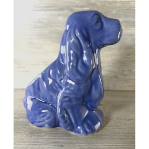 May include: A glossy, solid blue ceramic figurine of a sitting dog with long, textured ears and a smooth, rounded snout. The sculpture features a reflective finish and is depicted in a seated position, capturing the canine's upright posture.