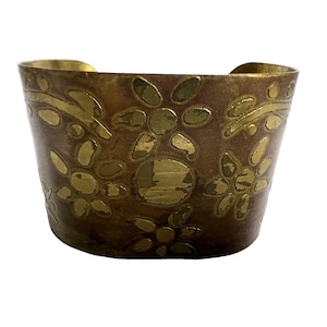 May include: A wide, brass-toned cuff bracelet with a floral design. The bracelet has a dark brown background with gold-colored floral cutouts. The edges of the bracelet are also gold-colored.