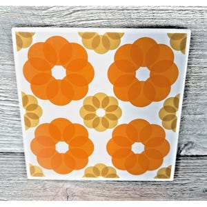 Op de afbeelding: Vierkante keramische tegel met een retro bloemenontwerp. De tegel heeft vier grote oranje bloemachtige vormen en kleinere geel-oranje bloemaccenten op een witte achtergrond. Het ontwerp is symmetrisch en gedurfd, met een vintage esthetiek.