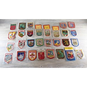 Puede incluir: Una colección de treinta y un parches de recuerdo bordados y coloridos, cada uno envuelto individualmente en plástico transparente. Los parches presentan diversas formas de escudos y emblemas, mostrando ilustraciones de ciudades, monumentos y símbolos regionales europeos.
