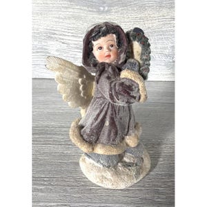 May include: A vintage angel figurine with a mauve dress, cream-colored wings, and a matching hood. The angel is holding a wreath and is standing on a snow-covered base. The figurine has a detailed face and is made of a textured material.