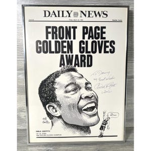 May include: A framed black and white newspaper front page from the Daily News, dated March 23, 1964, featuring a drawing of Emile Griffith and the headline "FRONT PAGE GOLDEN GLOVES AWARD." The image includes Griffith's signature and a small cartoon.