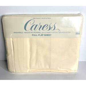 May include: A packaged full flat sheet in a cream color. The packaging has the word "Caress" in a script font, along with text indicating it is washable, needs no ironing, and is a 50% combed cotton/50% polyester blend.