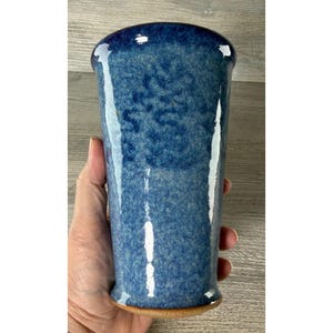 May include: A tall, cylindrical ceramic tumbler featuring a mottled blue glaze with varying shades of deep navy and lighter sky blue. The surface has a glossy, reflective finish with a smooth texture and a slightly flared rim at the top.