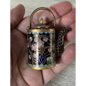 May include: A miniature metal teapot featuring black enamel with pink floral patterns, blue scalloped borders, and a gold-toned finish. The spout has a textured scale pattern, and the lid is topped with a small finial. It includes a thin wire handle.