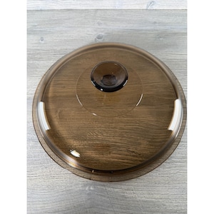 May include: A clear, brown-tinted glass lid with a dark brown knob. The lid is circular with a slightly raised rim and a central knob for easy handling. The glass has a smooth, reflective surface.