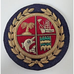 Op de afbeelding: Een ronde, marineblauwe embleem met een rood middenstuk verdeeld in vier secties. Elke sectie heeft een gouden geborduurd ontwerp, waaronder een palmboom, schip, dolfijn en gestileerde groene en gouden vormen. Het embleem is omcirkeld door een gouden lauwerkrans.