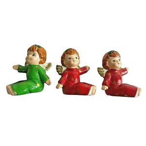 May include: Three ceramic angel figurines in green and red. Each angel has a smiling face, brown hair, and small gold wings. The angels are sitting with their arms outstretched, and are decorated with gold star accents.