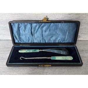 May include: A vintage set featuring a silver-toned shoehorn and a button hook, both equipped with marbled light green handles. These two tools are presented inside a rectangular black case lined with dark blue velvet and light blue satin.