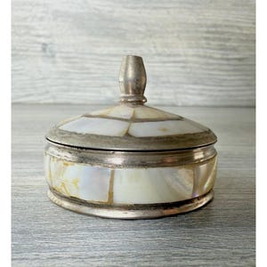 May include: A round lidded container featuring a metallic silver-toned frame and inlaid iridescent mother-of-pearl panels. The lid includes a central silver-toned knob handle, and the body displays a segmented pattern of white and cream shell.
