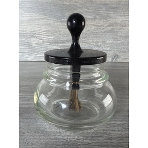May include: A clear glass jar with a black lid and handle. The lid has text on it. A brush extends from the lid into the jar. The jar is sitting on a gray surface.