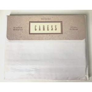 Op de afbeelding: Een verpakt twin plat laken. De verpakking is wit met een bruin bovenstuk. Het bovenstuk heeft de tekst "Twin Flat Sheet" en "CARESS". De verpakking vermeldt ook "No Iron Percale", "180 Thread Count", "50% Cotton" en "50% Polyester".