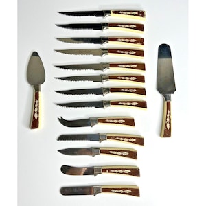 Puede incluir: Un juego de cuchillos de carne y utensilios para servir vintage. Los cuchillos tienen hojas dentadas y mangos marrones con detalles crema y diseños de hojas. También se incluye una espátula para pastel.