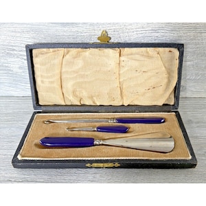 May include: A set of three metal grooming tools with deep blue handles, including a long button hook, a short button hook, and a shoehorn. The items are presented in a rectangular black case with a beige velvet lining and a cream fabric lid.