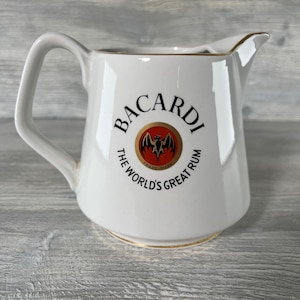 Può includere: Brocca in ceramica bianca con manico e beccuccio. La brocca presenta il logo "BACARDI" in nero con un emblema rosso e oro e la scritta "THE WORLD'S GREAT RUM". Il bordo e la base hanno un bordo dorato.