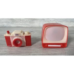 May include: Two miniature toys: a red and cream-colored camera and a red and cream-colored television. The camera has a red lens and a red button on top. The television has a white screen and a red base. Both toys are on a light gray surface.