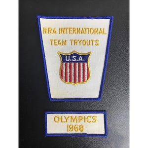 Op de afbeelding: Geborduurde patchset met de tekst "NRA INTERNATIONAL TEAM TRYOUTS" en "OLYMPICS 1968". De bovenste patch heeft een schild met "U.S.A." en rood-witte strepen. Beide patches hebben een witte achtergrond met blauwe randen.