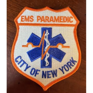 May include: A shield-shaped embroidered patch with an orange border. The white background features the text EMS PARAMEDIC in orange and CITY OF NEW YORK in blue. A blue Star of Life symbol is centered with an orange EKG line and a white staff.