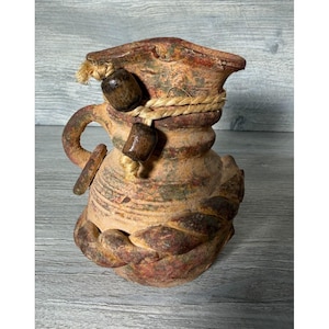 May include: A rustic, hand-crafted ceramic vase with a unique design. The vase features a textured, aged appearance with earthy tones of brown, red, and green. A rope and wooden beads adorn the neck, adding a touch of natural charm. The vase has a handle.