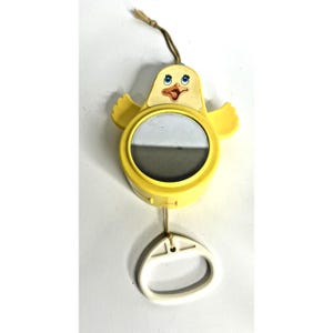 Op de afbeelding: Een vintage gele baby mobile in de vorm van een cartoon kuiken met een spiegel. Het kuiken heeft blauwe ogen, een oranje snavel en kleine gele vleugels. Een witte plastic ring hangt aan de onderkant en een bruin touwtje is aan de bovenkant bevestigd.