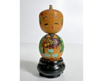 May include: A vintage wooden Japanese Kokeshi doll. The doll features a round head with painted facial features and a cylindrical body decorated with painted designs. It sits on a black, round base.