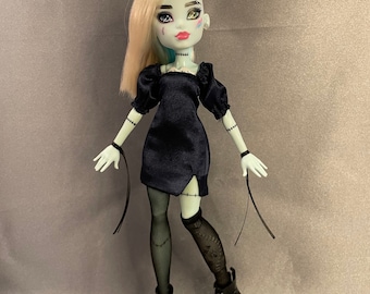 Puede incluir: Una muñeca de moda de piel azul con cabello rubio y azul que lleva un vestido negro con mangas abullonadas y botas negras. La muñeca tiene ojos verdes y azules y detalles cosidos en su rostro y brazos.