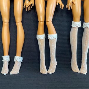 May include: Doll legs with white lace-trimmed socks. The socks are a sheer, dotted pattern. The doll legs are a tan color, and the background is a dark gray.