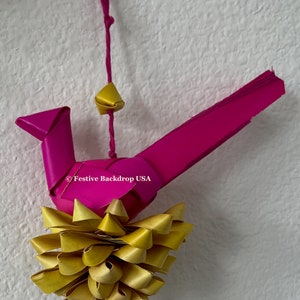 May include: A pink paper bird ornament with a yellow paper flower and a gold bell hanging from it. The ornament is hanging from a pink string.