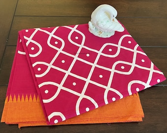 May include: A pink cotton saree with a white geometric pattern and an orange border. The saree is folded and displayed on a wooden surface.