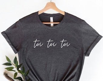 Puede incluir: Una camiseta gris oscuro con la frase "toi toi toi" escrita en blanco en la parte delantera.