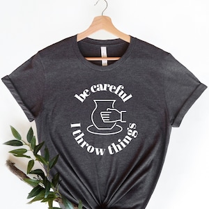 May include: A dark grey t-shirt with a white graphic that says "be careful I throw things" and a silhouette of a hand throwing clay on a pottery wheel.