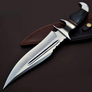 May include: A silver and black hunting knife with a serrated blade and a black handle with gold accents. The knife is resting on a brown leather sheath.